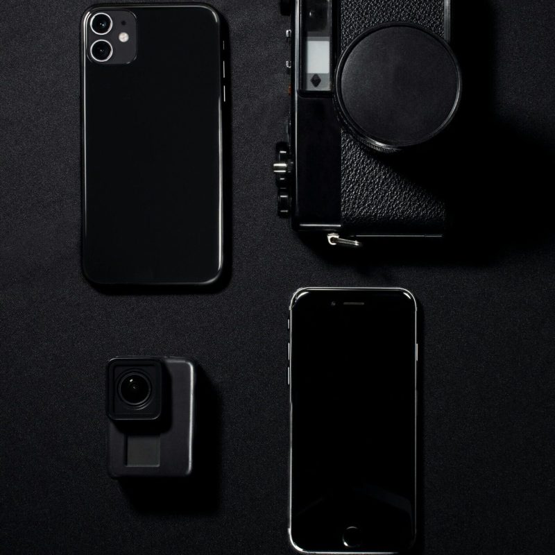 Minimalist flat lay of black gadgets including phones and a camera against a dark background.